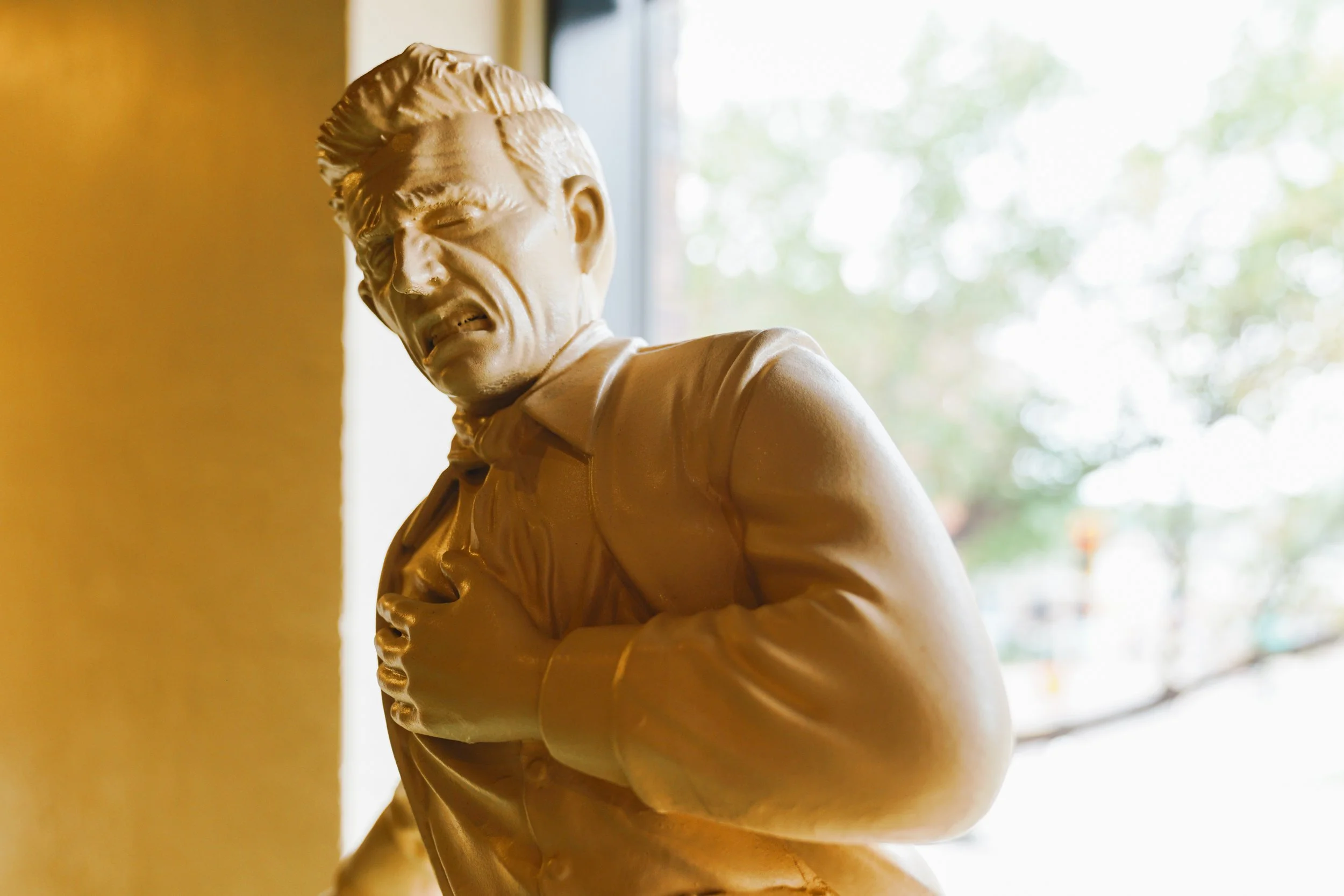 Close-up of a golden statue of a man with an anxious or pained expression, set near a window with blurred green trees outside.