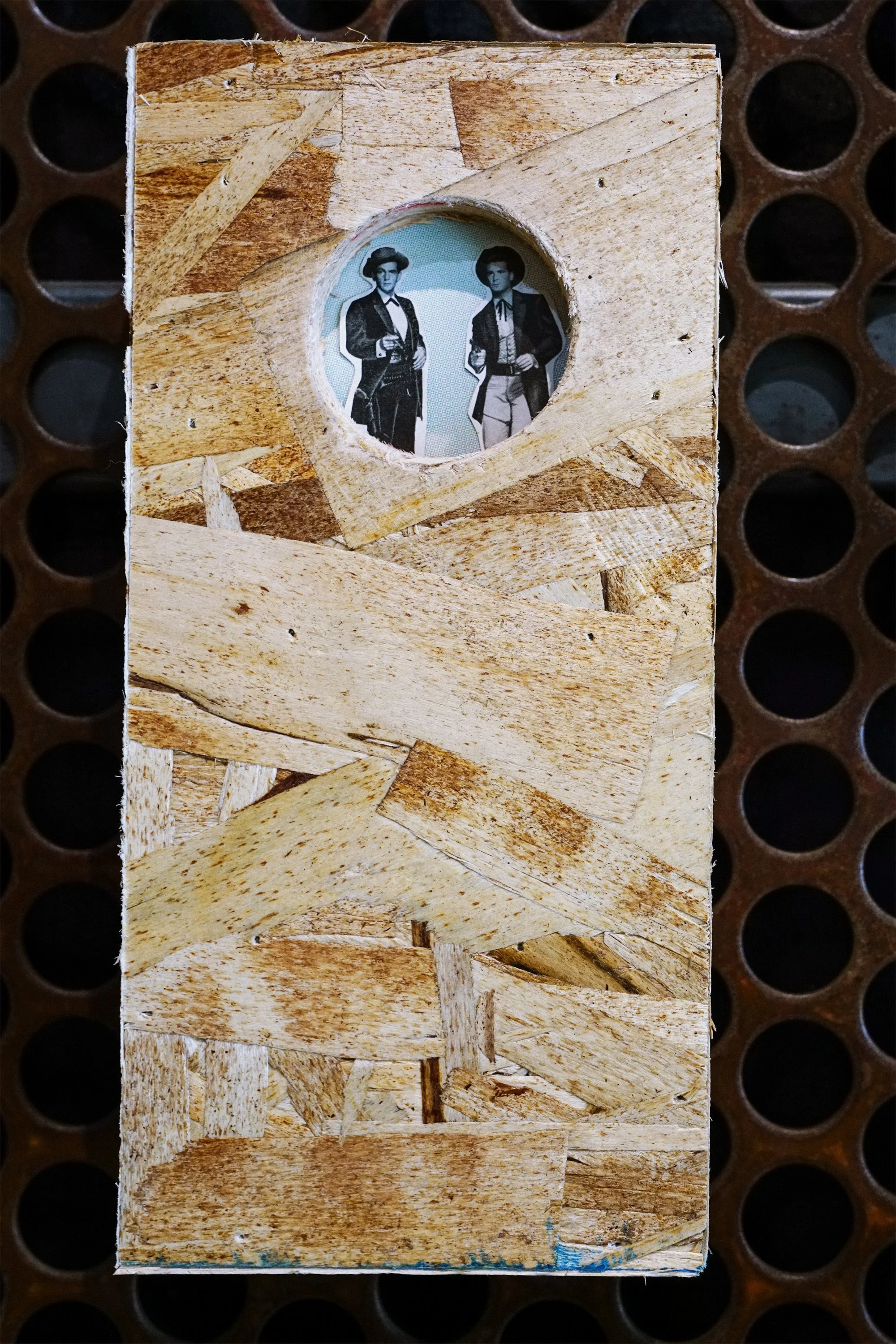 A vintage photo of two women dressed in early 20th-century clothing, seen through a circular cutout in a wooden board. The wooden board has a rough, patchwork appearance. The background is a metal surface with circular holes.