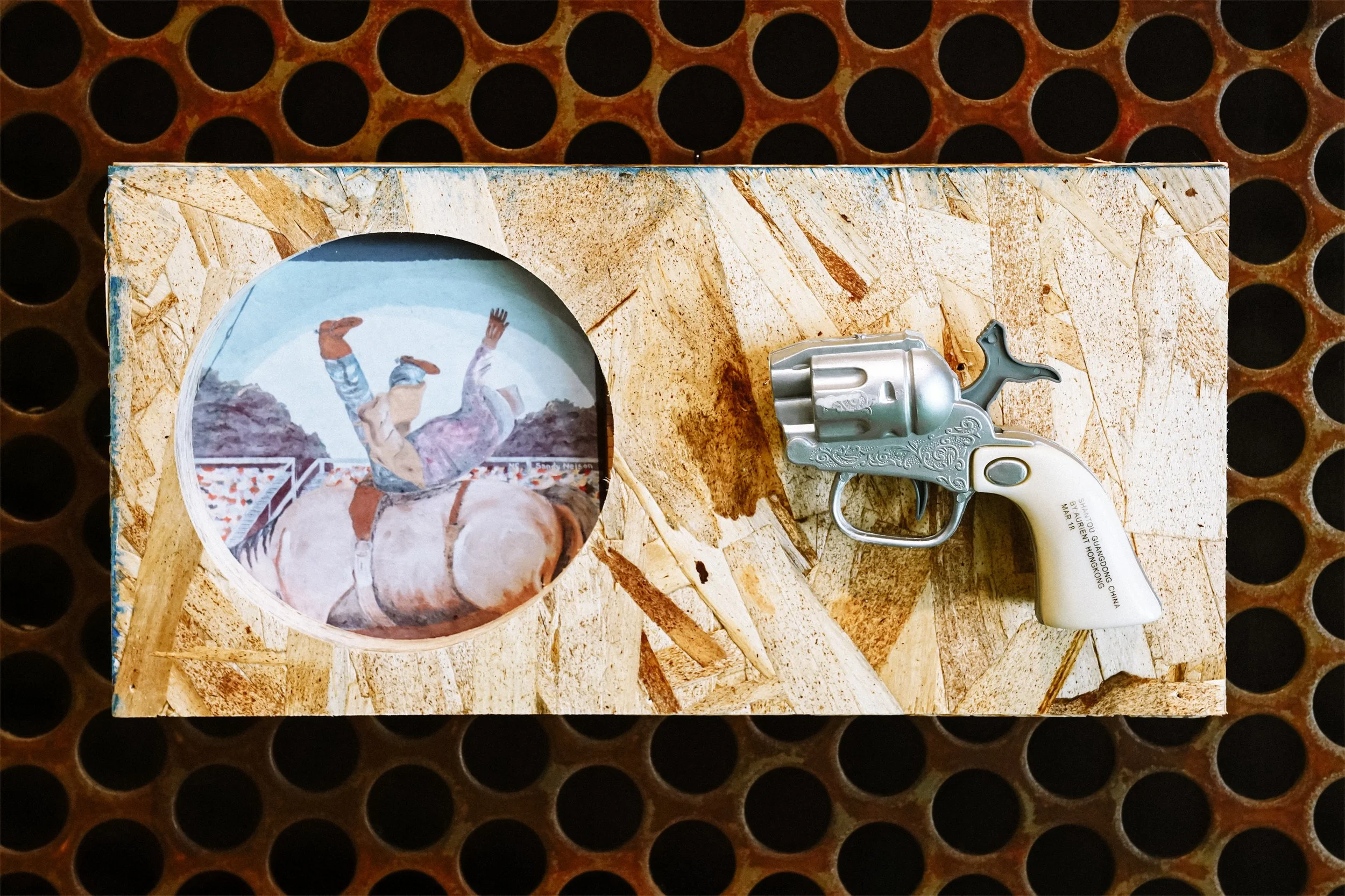 A decorative box with a cowboy scene painting and an antique toy revolver placed on a wooden surface with circular hole pattern background.