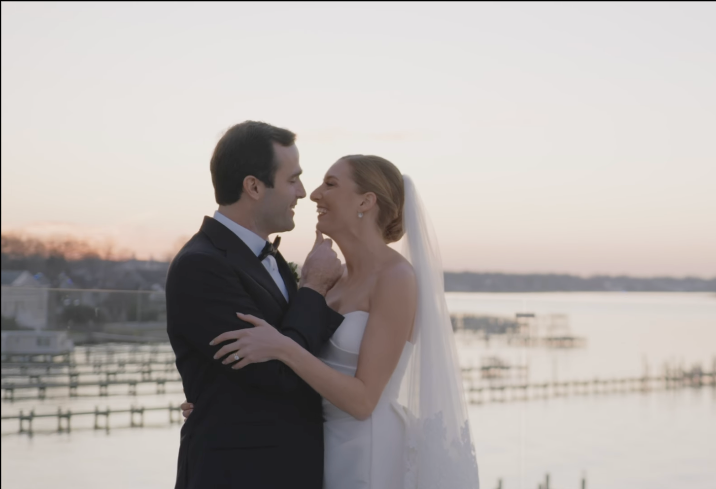 A bride and groom share a tender moment by the water at sunset, smiling and touching noses.