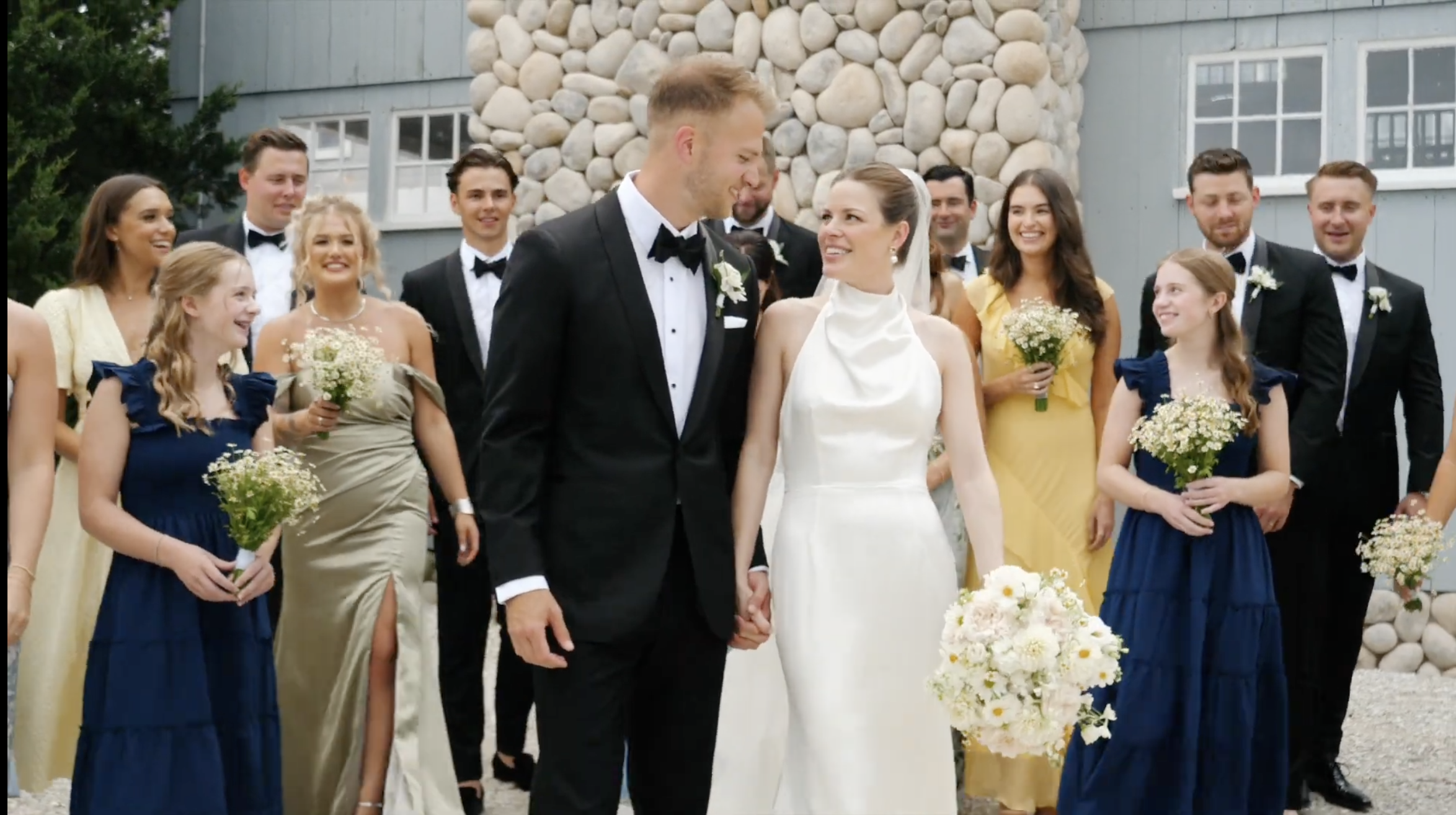 A wedding celebration with a bride and groom holding hands surrounded by bridesmaids and groomsmen outside a building with a stone foundation.