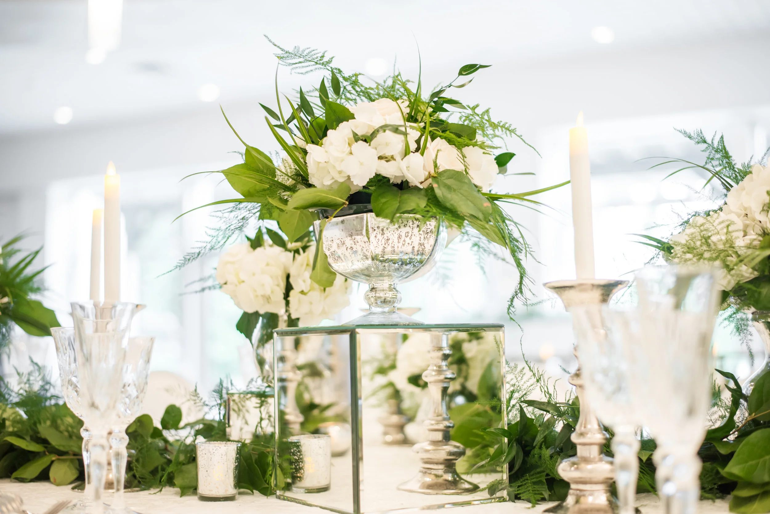 Lyric Flowers elevated the Ballroom design with an abundance of white floral and greenery, arranged in low mercury dish centerpieces, raised in glass urns, and laced through chandeliers.