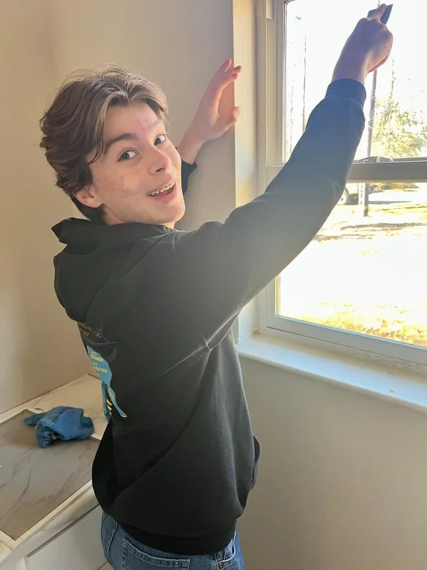 A young boy in a black hoodie and jeans standing near a window, smiling and reaching up to touch the window frame while holding a small object in his other hand, with sunlight outside.
