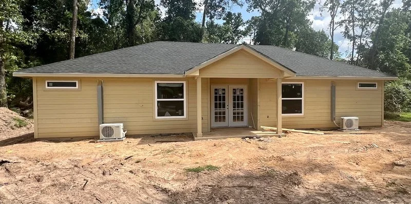 New beige house with front porch, surrounded by trees and cleared yard, with air conditioning units outside.