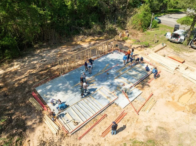 Construction workers building the foundation of a structure on a cleared lot with wooden framing and concrete slab, surrounded by trees and nearby vehicles.