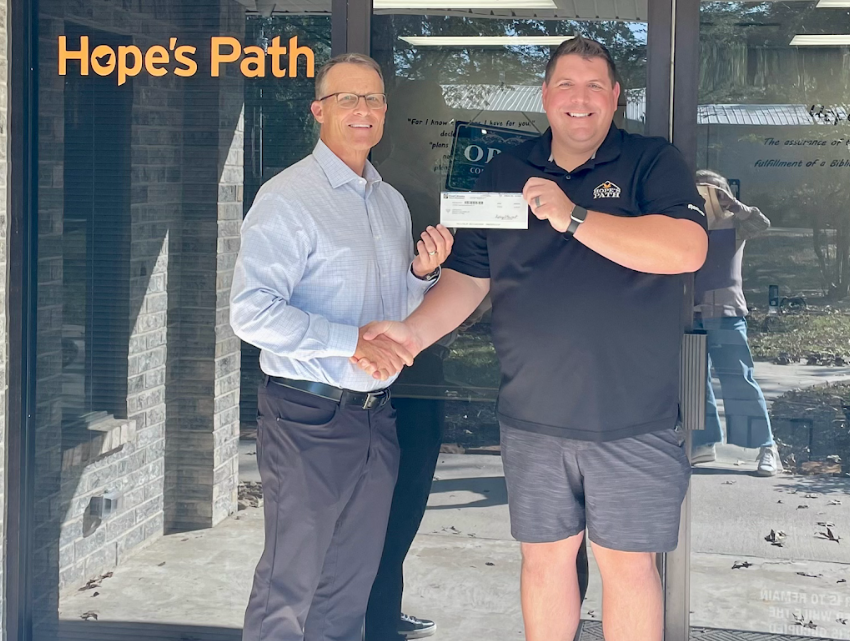 Two men shaking hands outside a building with a sign that reads 'Hope's Path.' One man is holding a check and both are smiling.