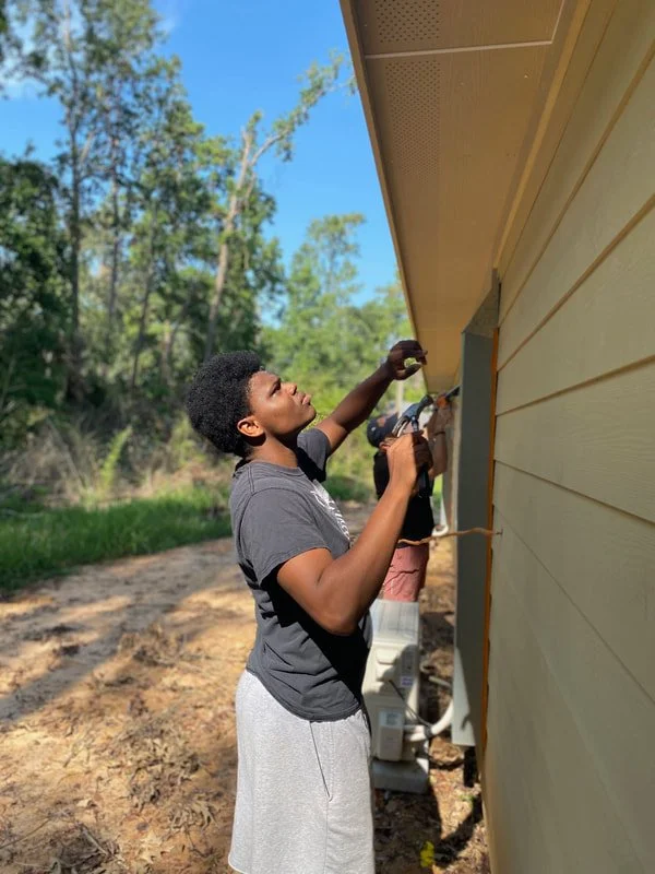 Young boy working on a house's exterior with a power drill, in a sunny outdoor setting surrounded by trees.