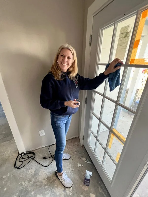 A woman cleaning a glass door with a cloth, holding a phone, smiling, wearing a navy hoodie, jeans, and white sneakers, in a bright room with a nearby electrical outlet and cleaning spray on the floor.