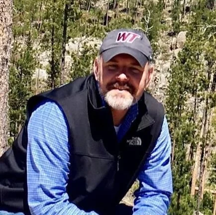 A man wearing a gray baseball cap with 'WT' on it, a blue checkered shirt, and a black vest outdoors in a forested area.