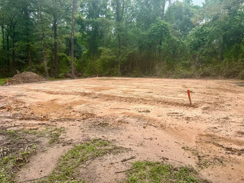 A cleared and leveled construction site surrounded by green trees, with orange markers in the dirt.