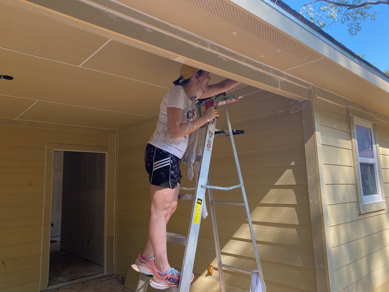 A woman standing on a ladder and working on the soffit of a house under a clear blue sky.