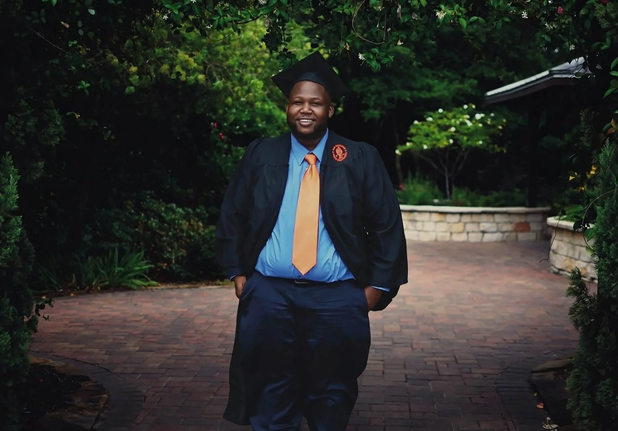 A man in a graduation cap and gown smiling as he walks outdoors on a brick pathway with greenery and bushes in the background.