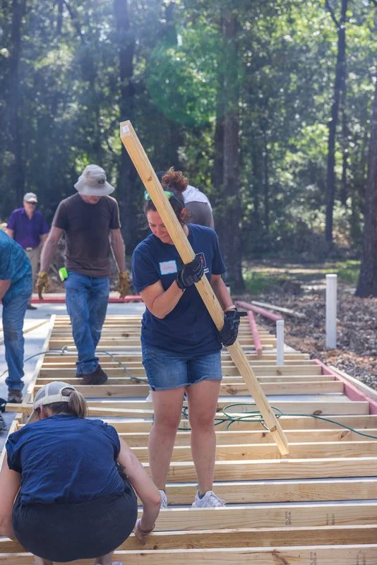People working on a construction site outdoors, assembling wooden framework for a structure surrounded by trees.