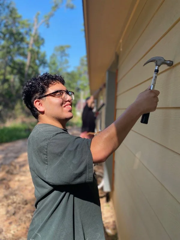 Young man wearing glasses and a dark t-shirt hammering nails into the siding of a house with another person working in the background on a sunny day.
