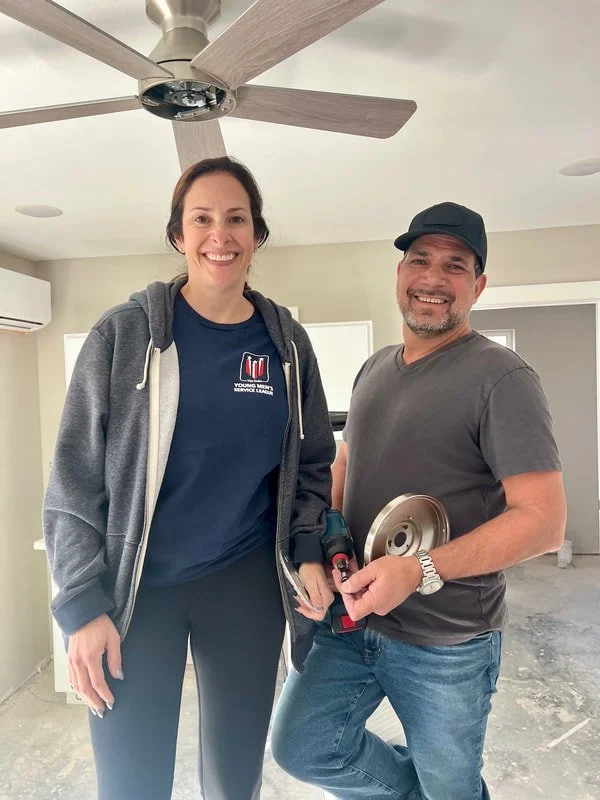 A woman and a man standing inside a room with a ceiling fan above them. The woman is smiling and wearing a navy T-shirt and a gray zip-up hoodie. The man is smiling, holding a power tool, and wearing a dark cap, gray T-shirt, and jeans.