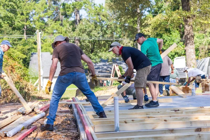 Group of people working together building a wooden structure outdoors, surrounded by trees and construction materials.