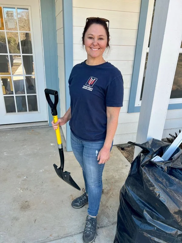A woman with brown hair tied back, wearing a navy blue T-shirt and jeans, holding a shovel, standing outside a house with white siding and a glass door.