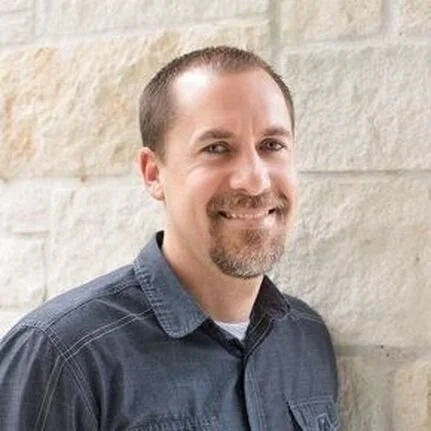 Portrait of a smiling man with short hair and a beard, wearing a dark denim shirt, standing against a light-colored brick wall.