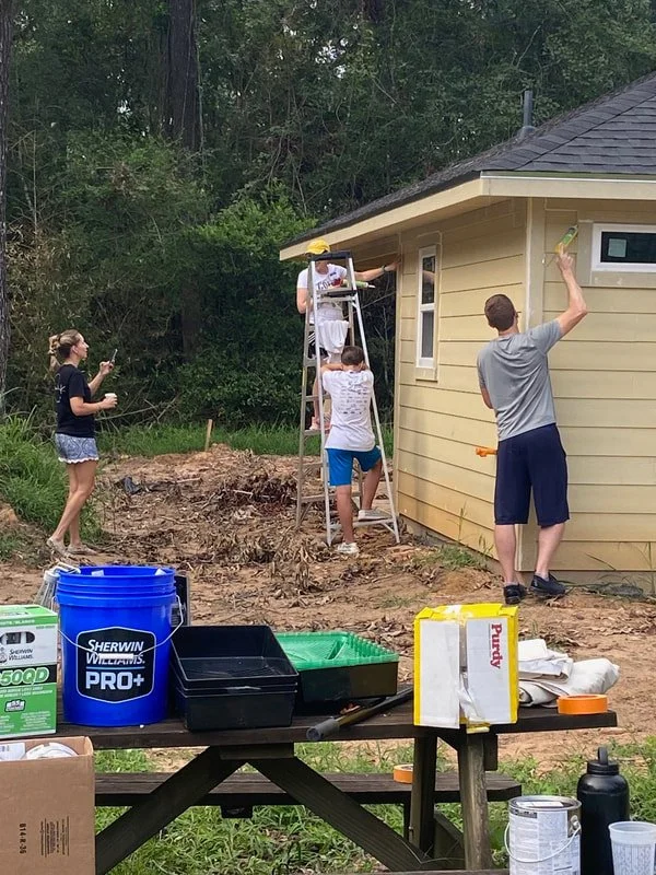 A group of five people working on a house exterior, with one person on a ladder painting, others painting the house or taking a photo, in a backyard with trees.