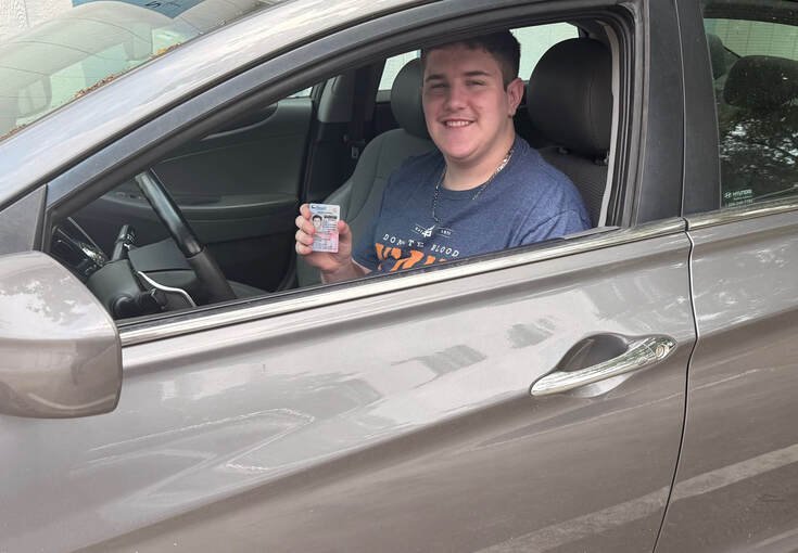 A young man sitting in the driver's seat of a silver car, smiling, holding an ID card, in an outdoor parking lot.