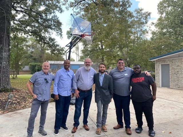 Six men standing in a row outdoors on a driveway, with trees and a house in the background. A basketball hoop is visible above them.