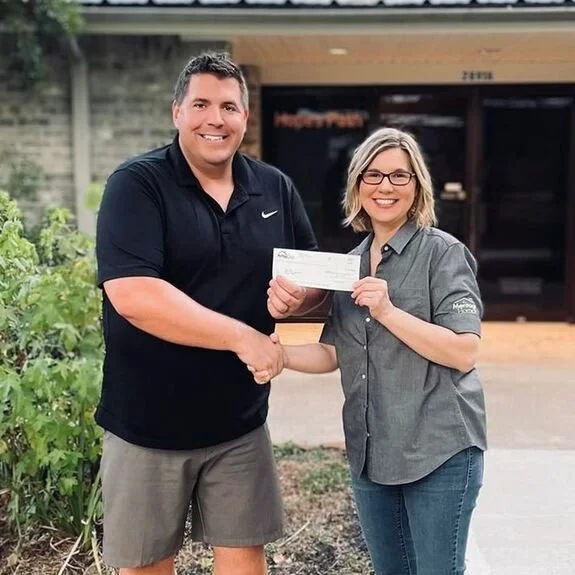 A man and woman shaking hands outdoors, with the woman handing the man a check. The man is wearing a black Polo shirt and khaki shorts, while the woman is wearing glasses and a gray button-up shirt.