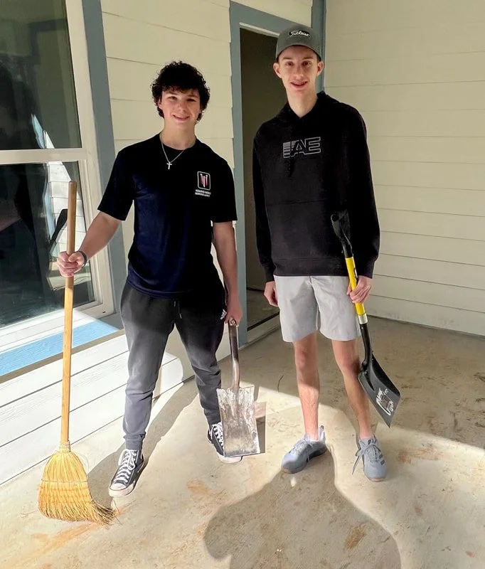 Two young men standing on a porch with cleaning tools, smiling at the camera. One holds a broom and a dustpan, the other a shovel.