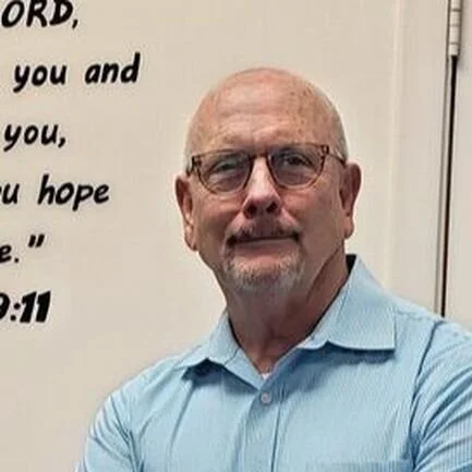 A middle-aged man with glasses and a beard, wearing a light blue dress shirt, standing in front of a white wall with a partial religious quote.