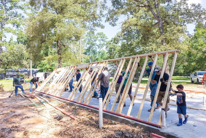 Group of people, including children, working together to build a wooden wall frame outdoors on a sunny day, surrounded by trees and grass.