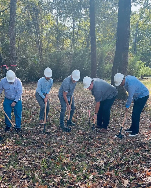 Five individuals wearing white hard hats and casual clothing are planting trees or shrubs in a wooded area, using shovels.