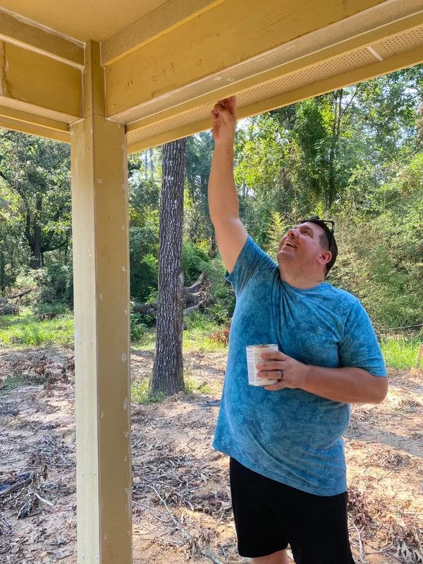 A man in a blue t-shirt and black shorts painting the underside of a porch ceiling with a small paintbrush, holding a container of paint, outdoors in a wooded area.