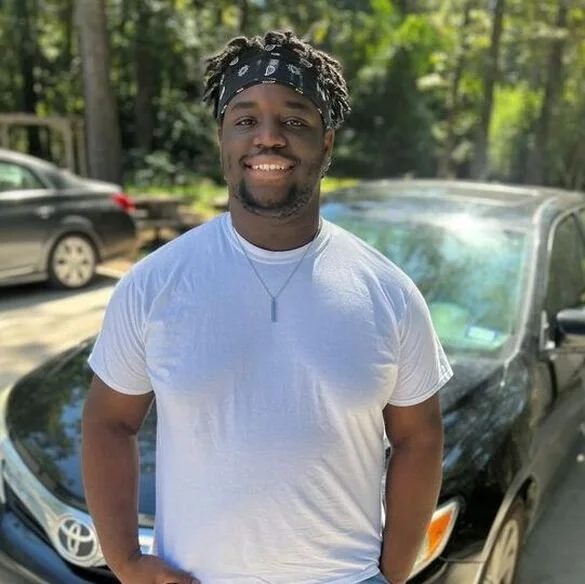 A smiling young man wearing a white t-shirt and a black bandana with white patterns, standing outdoors in front of a black car with trees in the background.