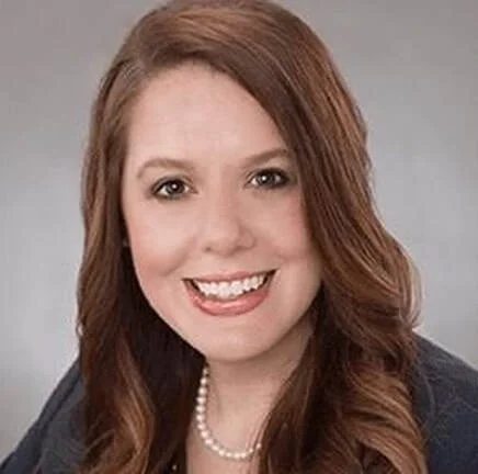 A smiling woman with long, wavy auburn hair, wearing a pearl necklace and a dark blazer against a neutral background.