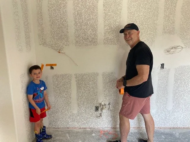 A young boy and an adult man standing inside a room under construction. The boy is wearing a blue T-shirt, red shorts, and boots, holding a yellow tool. The man is wearing a black cap, black T-shirt, and pink shorts, holding a measuring tape, smiling at the camera.