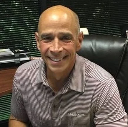 A smiling man sitting in an office chair with blinds and a desk behind him.