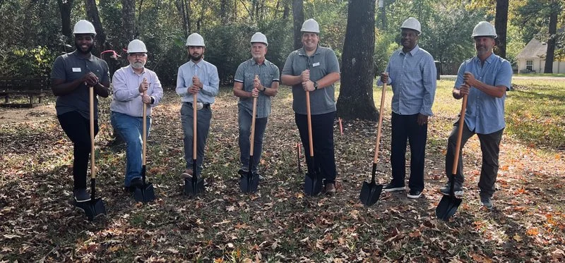 Seven men wearing hard hats standing outdoors in a line, holding shovels, in a wooded area with fallen leaves on the ground.
