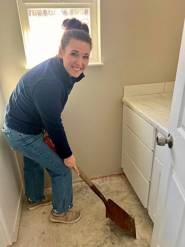 A woman with a bun hairstyle wearing a dark jacket and jeans, smiling and doing floor work with a pry bar in a small room with a window, white cabinet, and beige wall.