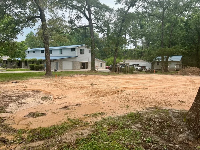 An empty plot of land with reddish dirt, surrounded by trees and residential houses in the background.