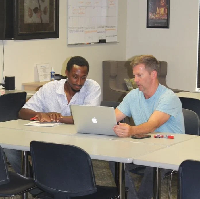 Two men sitting at a conference table looking at a laptop in an office setting.