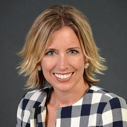 Headshot of a smiling woman with shoulder-length blonde hair, wearing a checked blazer and pearl earrings, against a gray background.