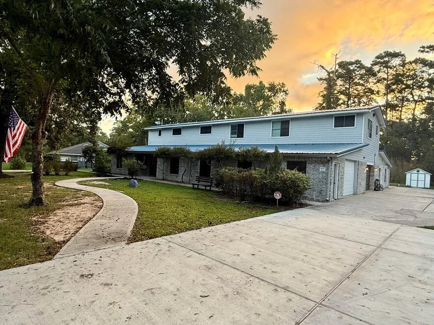 A two-story house with a driveway and a curved walkway leading to the front porch, surrounded by trees and bushes, under a colorful sunset sky.