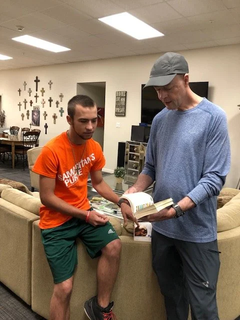 Two men standing by a beige couch, looking at an open book. One man is wearing an orange T-shirt and green shorts, and the other man is wearing a gray cap and a blue long-sleeve shirt.