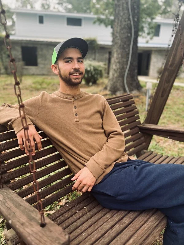 A young man sitting on a wooden swing in a backyard, smiling at the camera. He is wearing a brown long-sleeve shirt, dark pants, and a baseball cap. In the background, there are trees and a house with a stone base and light-colored upper walls.