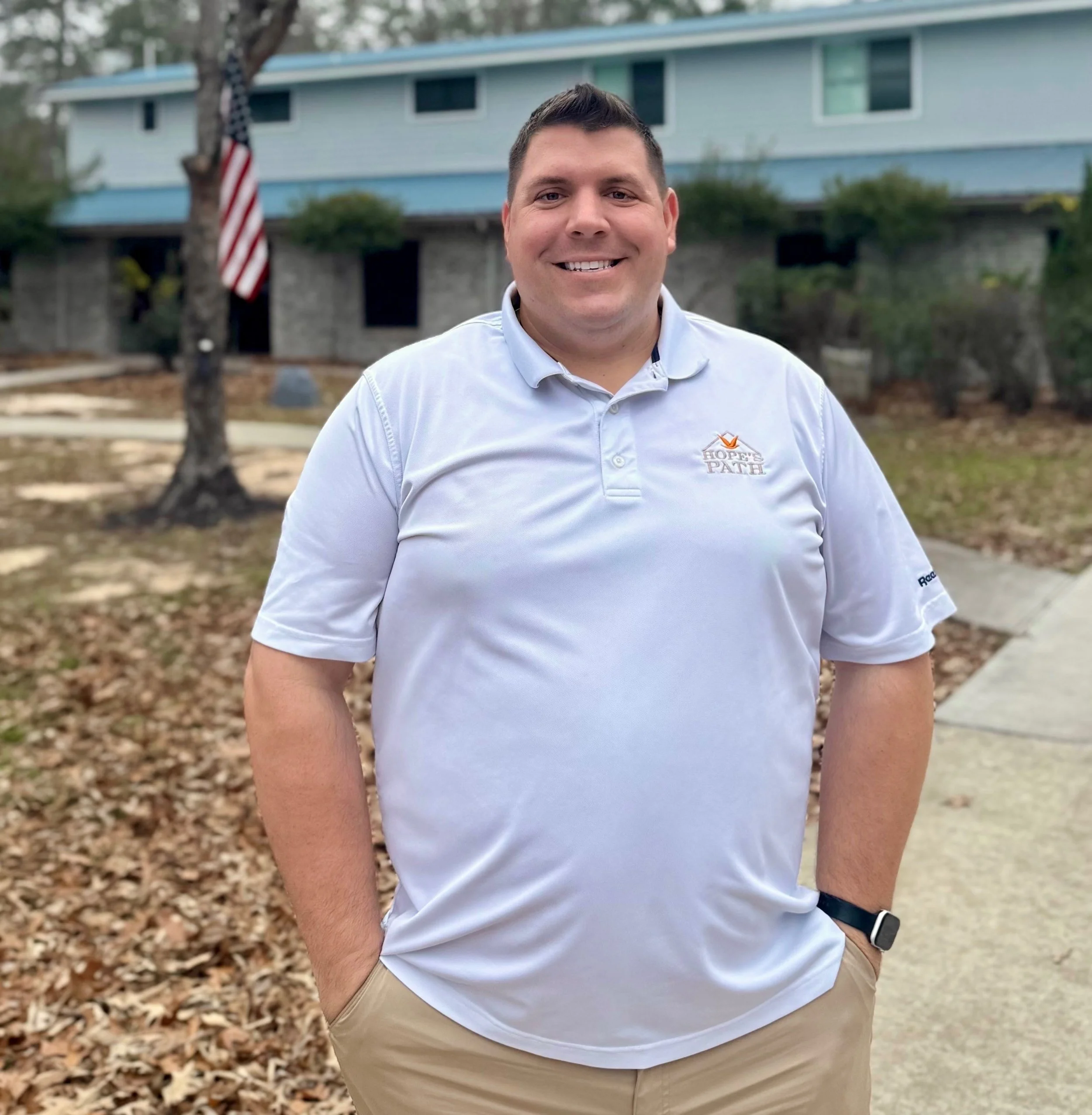 A smiling man standing outdoors on a sidewalk, wearing a white polo shirt and khaki pants, with a building, trees, and an American flag in the background.