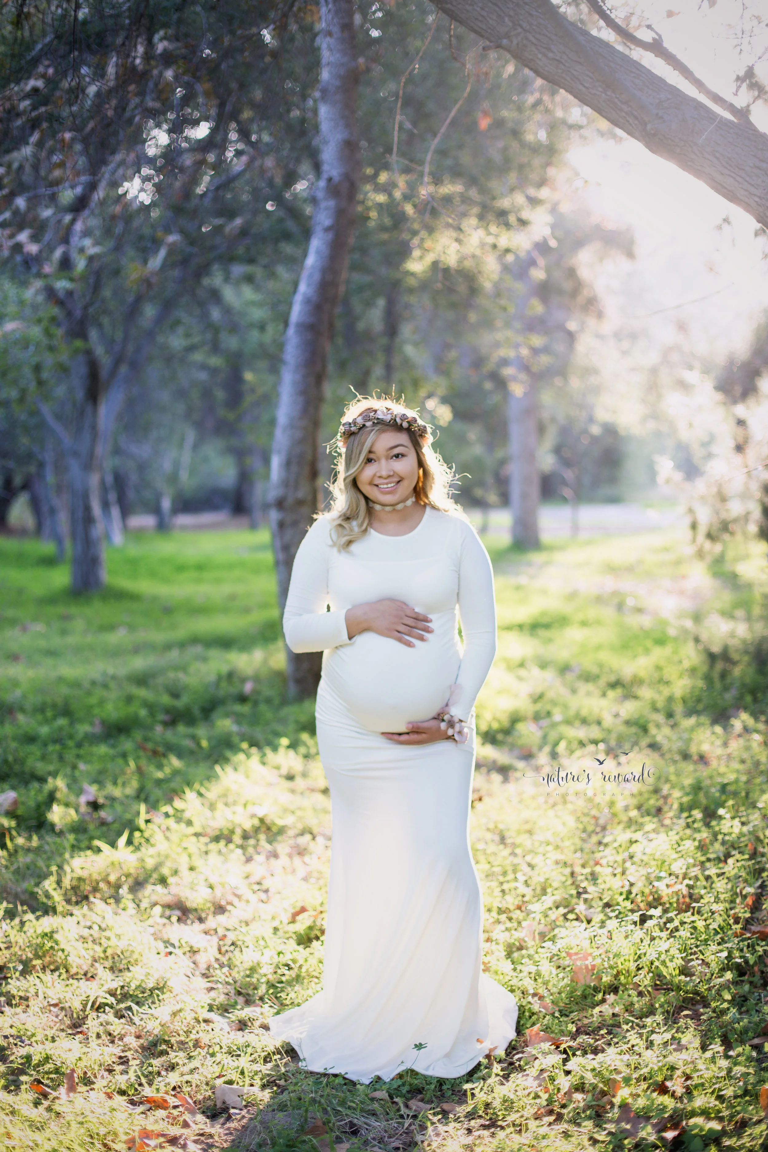 Mother to be in a white beautiful gown and flower crown smiles under a ray of light in this gorgeous maternity session portrait. 