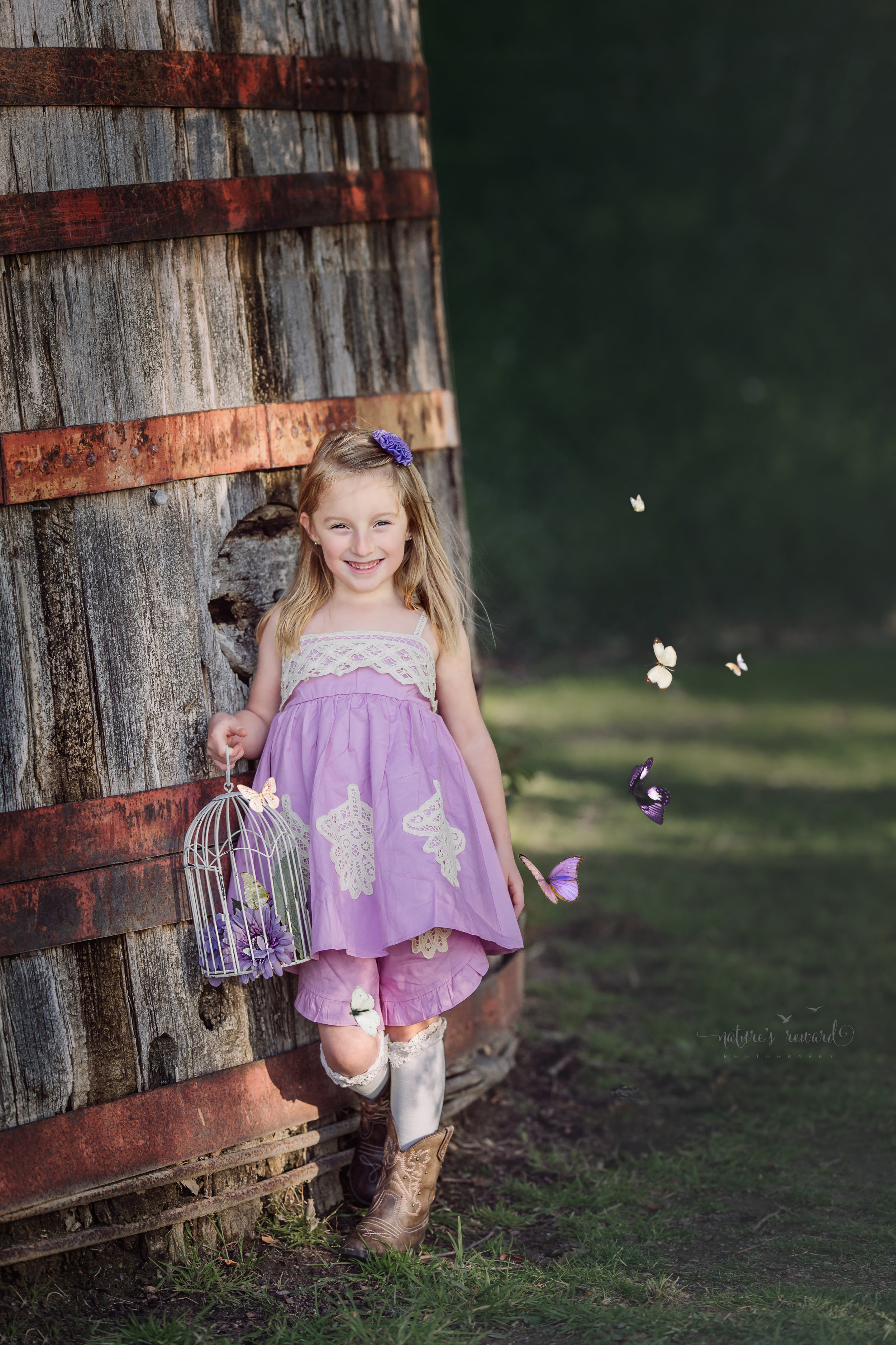 Butterflies fly around this sweet young girl holding a bird cage and smiling with joy wearing a purple dress and cowboy boots in this lovely spring portrait