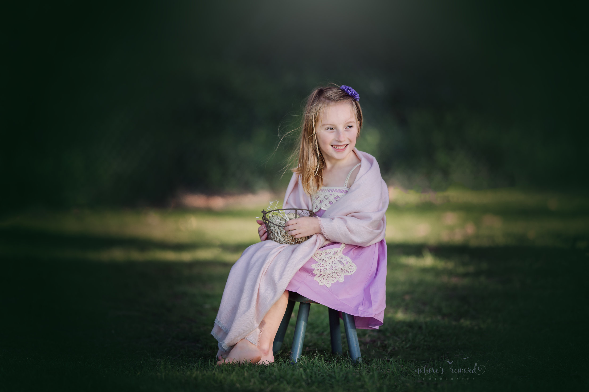 Gorgeous Spring portrait of a young girl in a purple dress sitting on a green stool wrapped in a soft pink scarf holding a basket of eggs filled with joy.