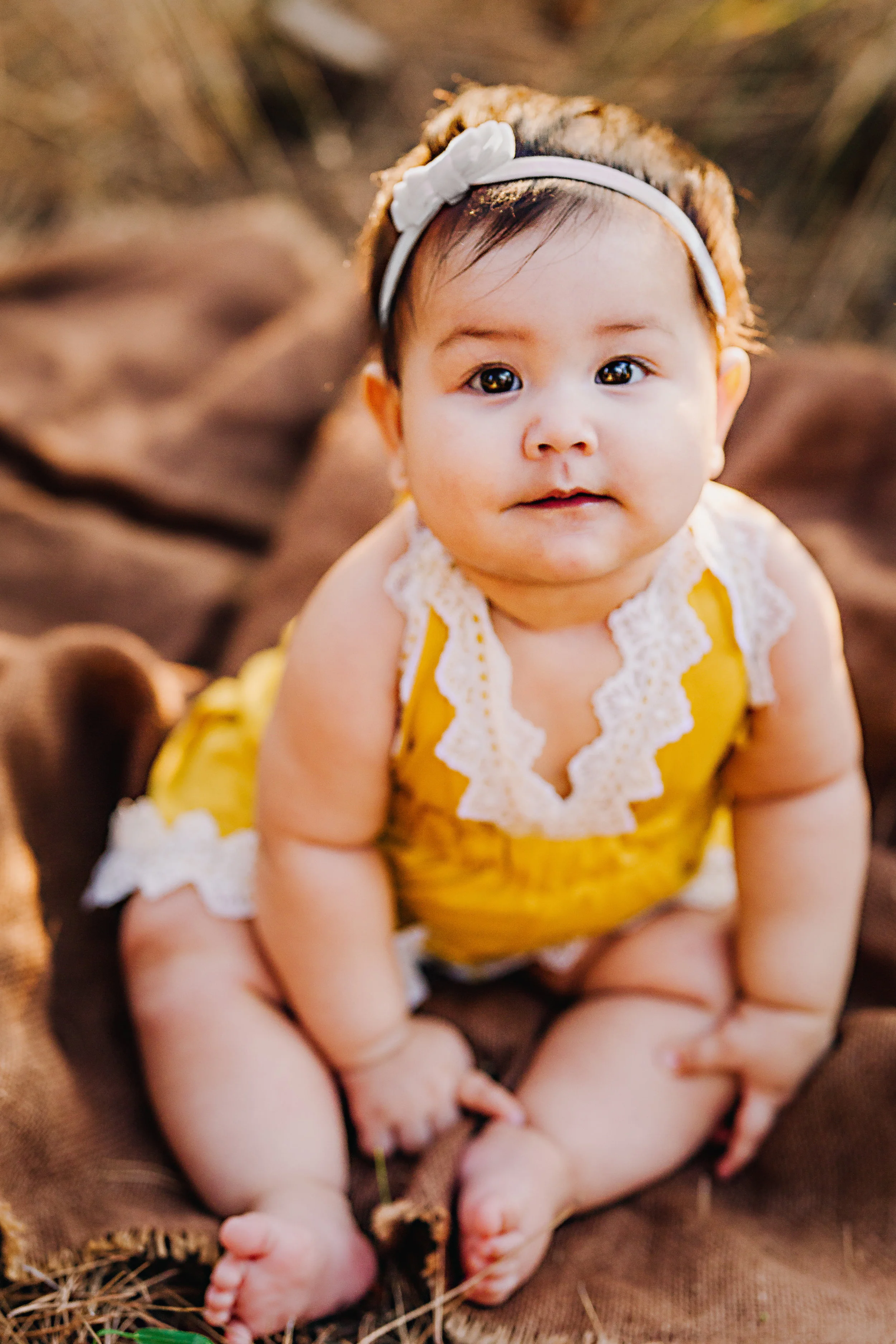Lovely baby in a yellow and white lace romper in the tall yellow reeds wearing a white hair bow showing off her baby rolls in this sitter portrait by Nature's Reward Photography