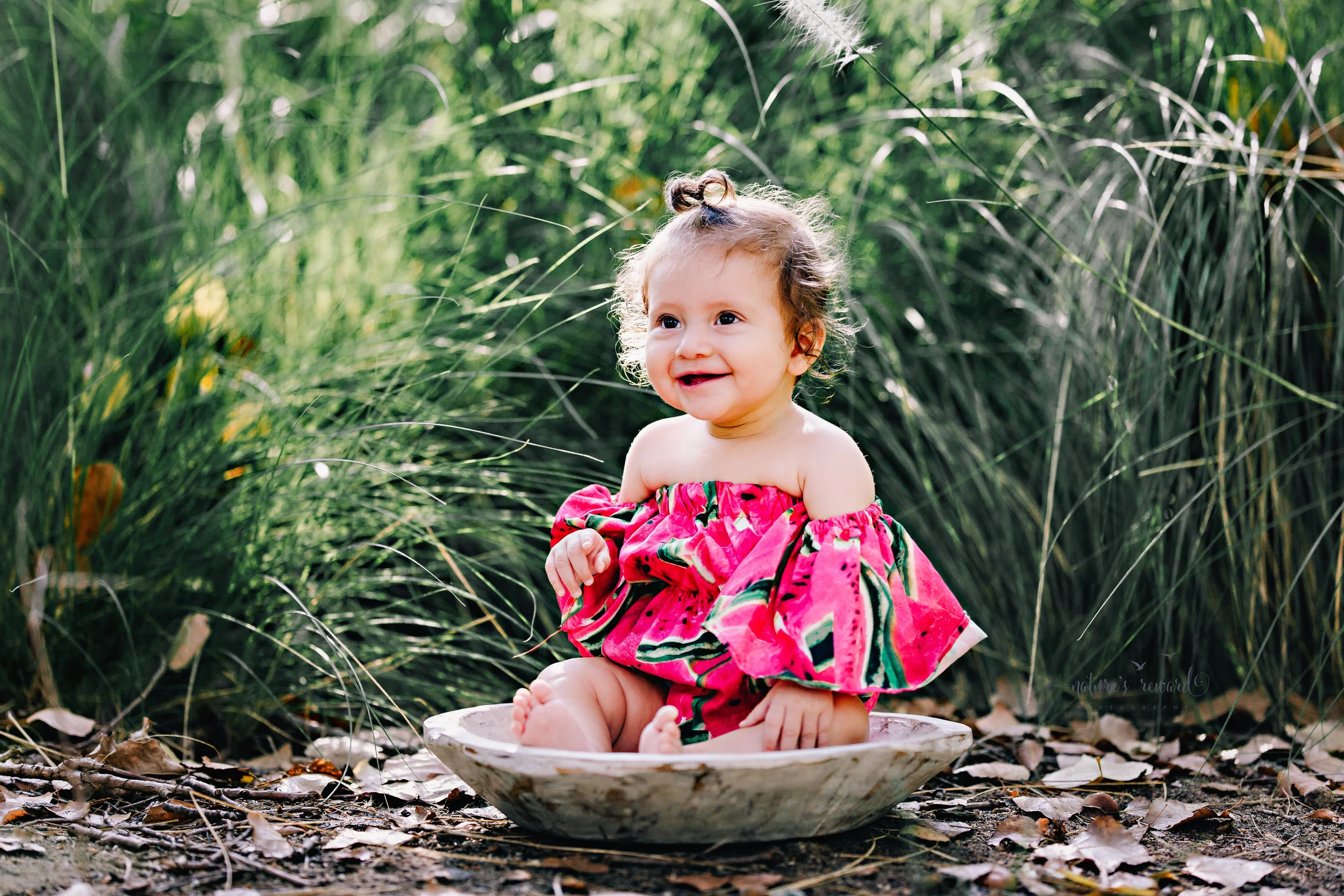 Gorgeous Baby girl in a watermelon off the shoulder romper sitting in front of the long reeds of grass during her sitter session in this portrait session by Nature's Reward Photography