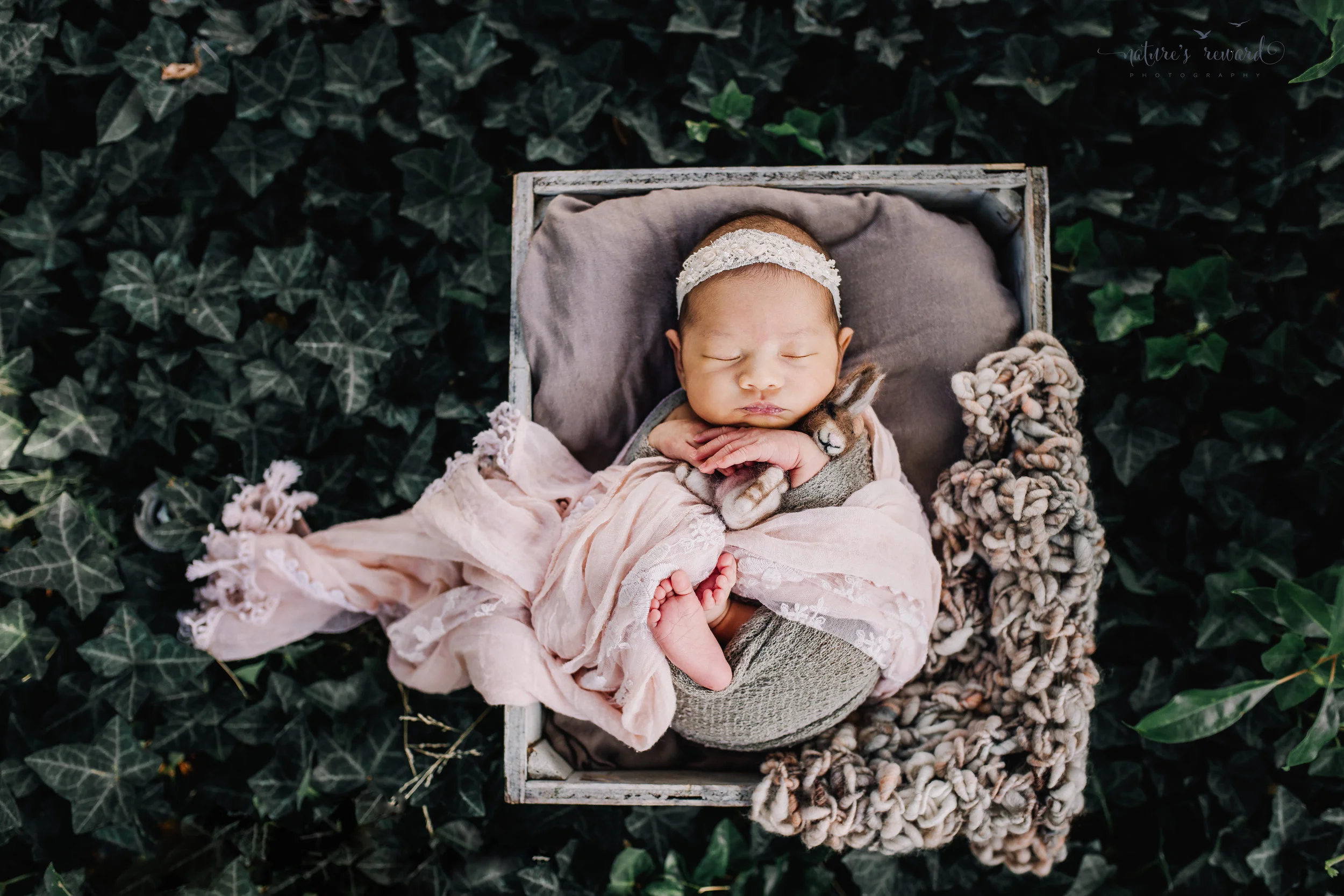 Gorgeous baby newborn girl swaddled in pink wearing a white tie back and in a garden surrounded by lush greens in this portrait by Nature’s Reward Photography